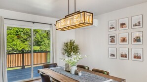 Un espace repas comprenant une table en bois, des chaises, un luminaire suspendu et une vue sur une terrasse en bois et la verdure environnante.