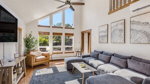 Un vaste salon comprenant un grand canapé d’angle gris, une table basse en bois et un téléviseur fixé au mur. De nombreuses fenêtres laissent entrer la lumière naturelle, et un ventilateur de plafond est visible.