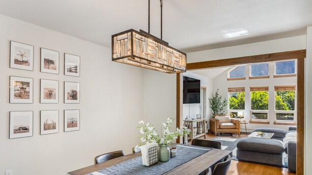 Un coin repas avec une table en bois, des chaises et un luminaire suspendu. Des tableaux encadrés ornent le mur, et on aperçoit le salon à travers une porte ouverte.