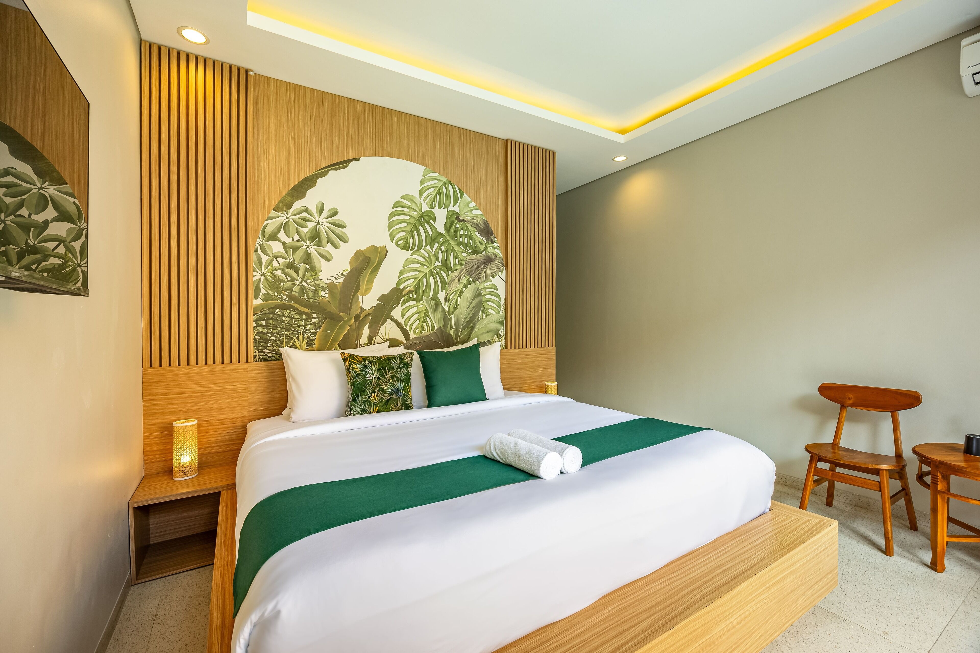 A modern hotel room with a wooden headboard, a large bed with green bedding, a nightstand with a lamp, and a small wooden table with a chair.