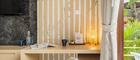 A room with a wooden desk, a chair, a television, a wall-mounted light, and a view of greenery outside.