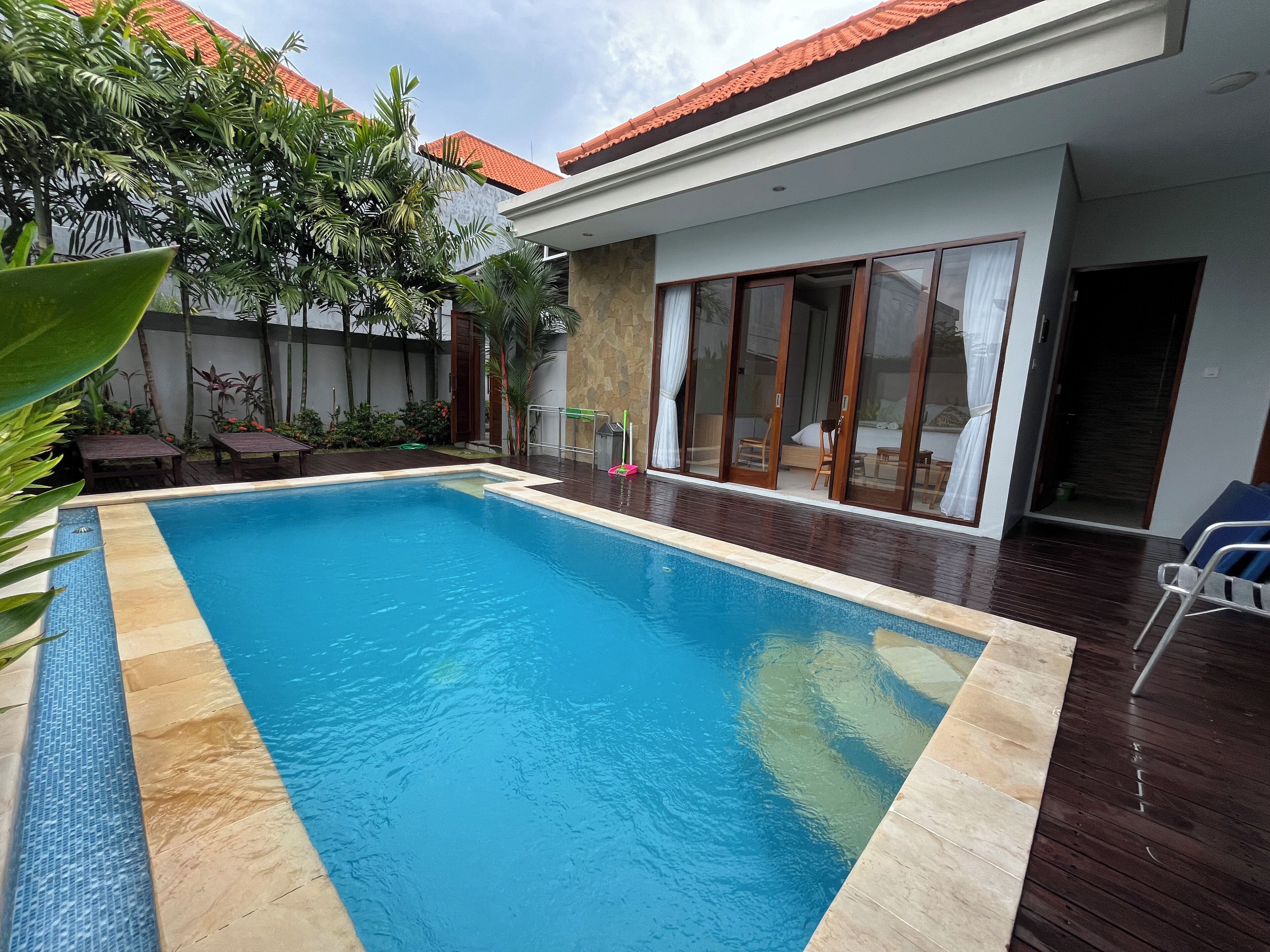 En swimmingpool med træterrasse, omgivet af beplantning og et hus med store glasdøre.