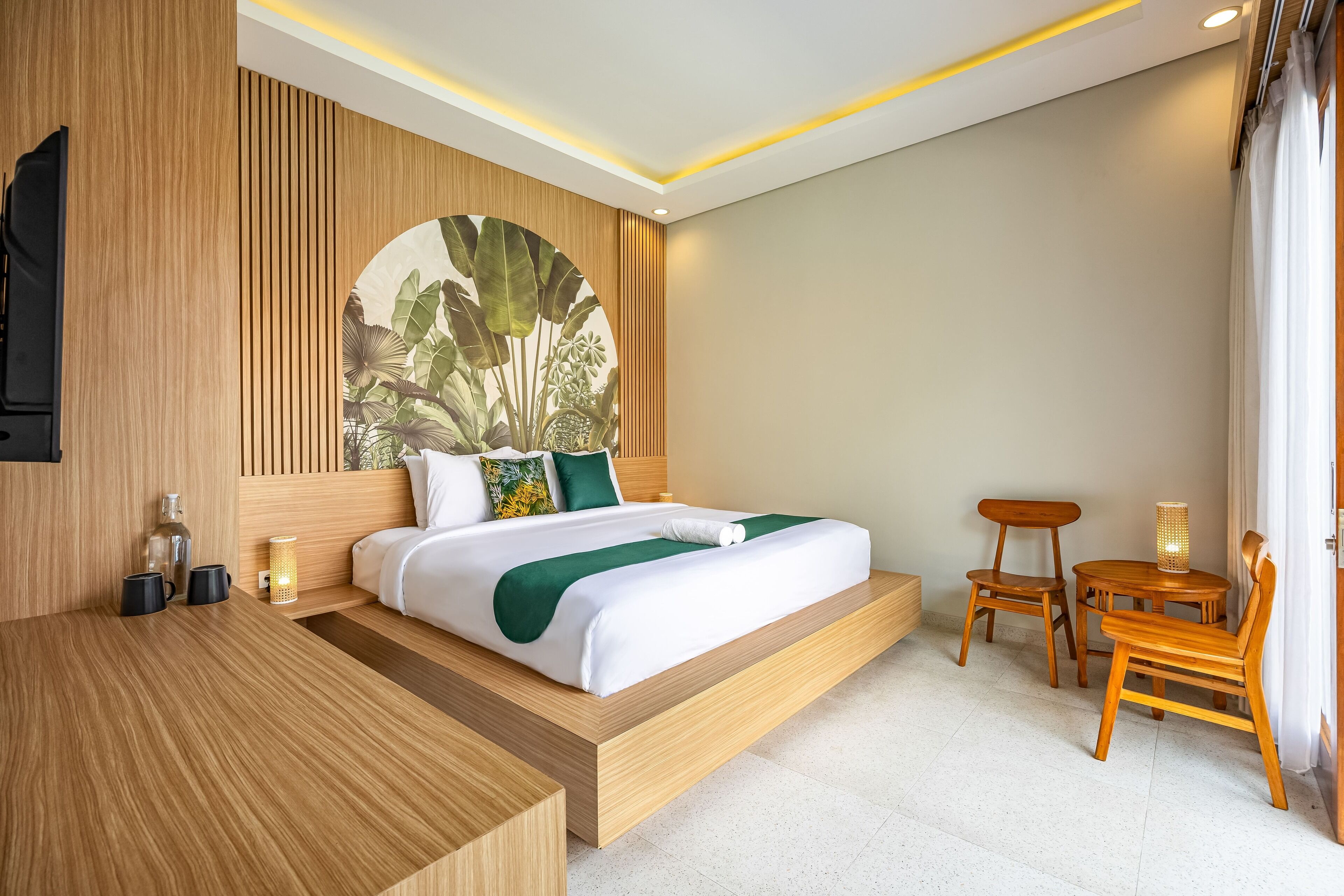 A modern hotel room with a wooden headboard, a large bed with white bedding and a green throw, a wooden side table, a small wooden chair, and a round wooden table.
