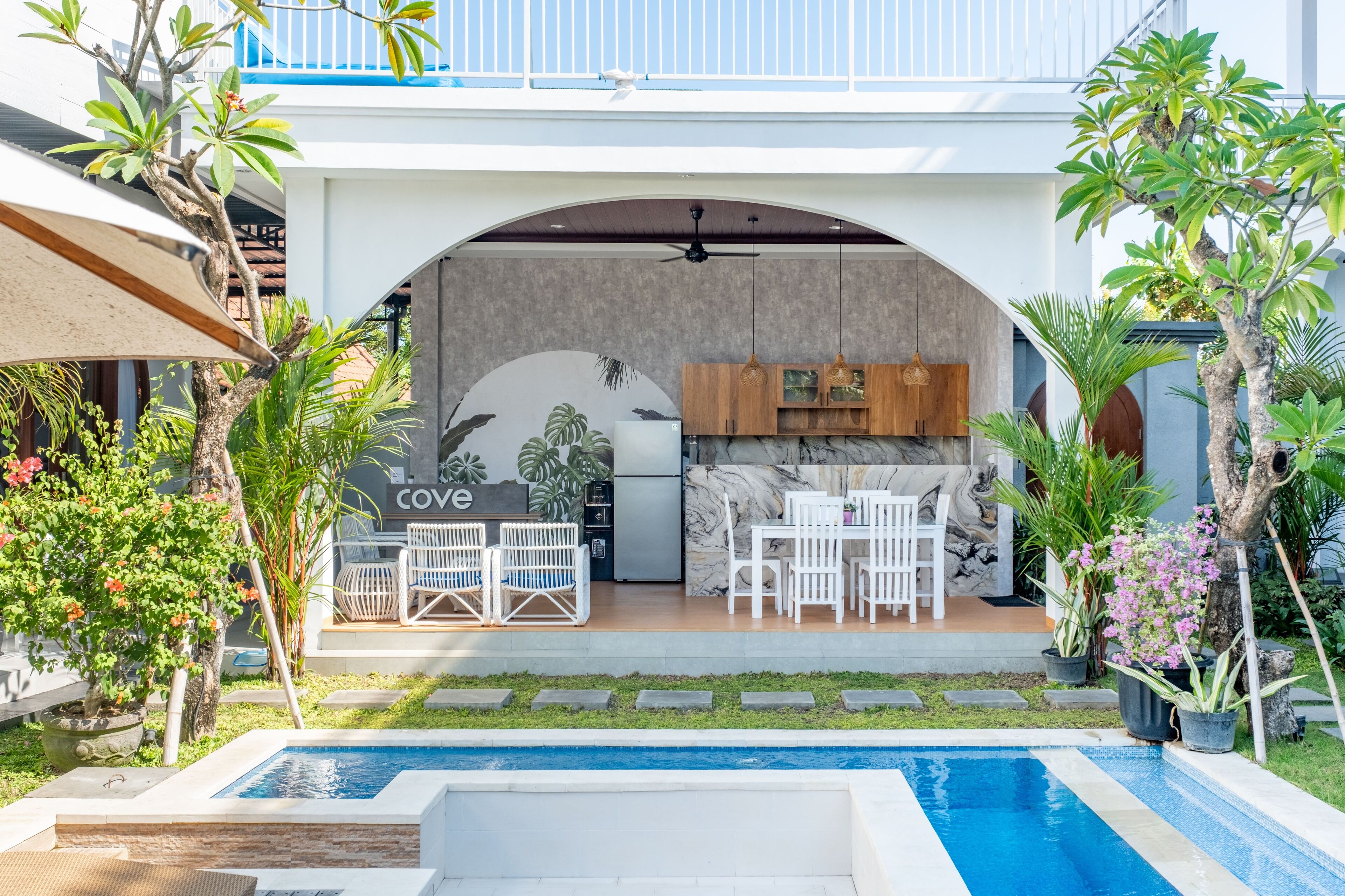 Un espace cuisine extérieur moderne, avec une piscine, entouré de verdure et agrémenté de mobilier blanc.