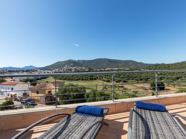 A balcony with two sun loungers, a view of a town and mountains in the distance.