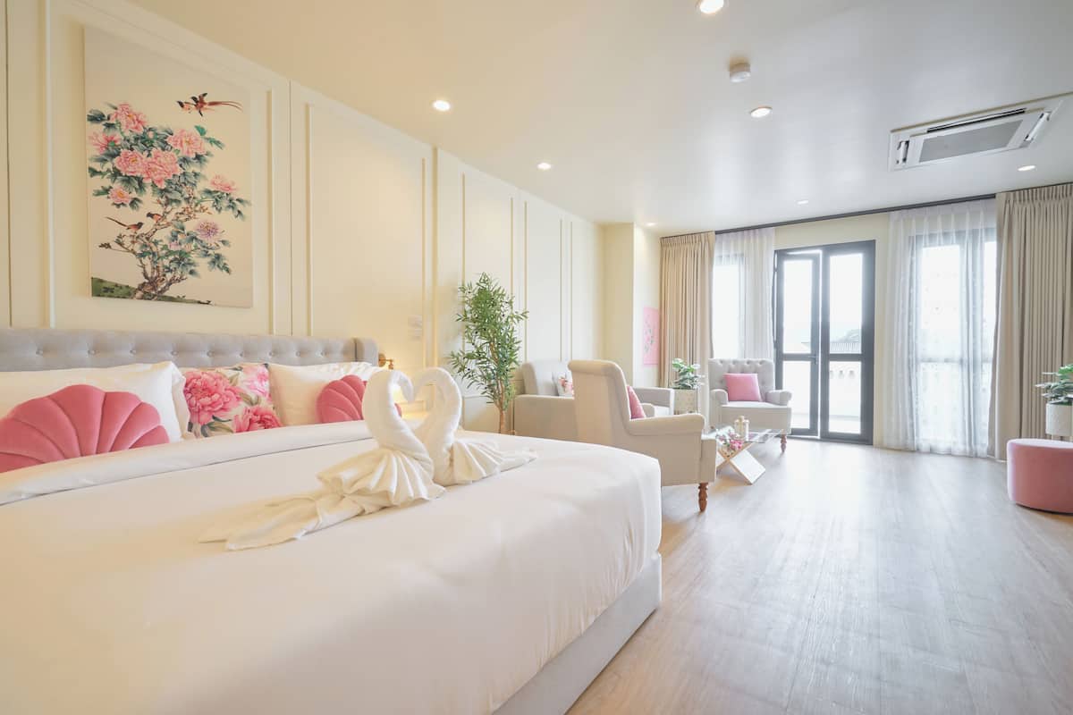 Habitación de hotel con una cama grande, una zona de estar con una silla y una mesita, y un ventanal con cortinas.
