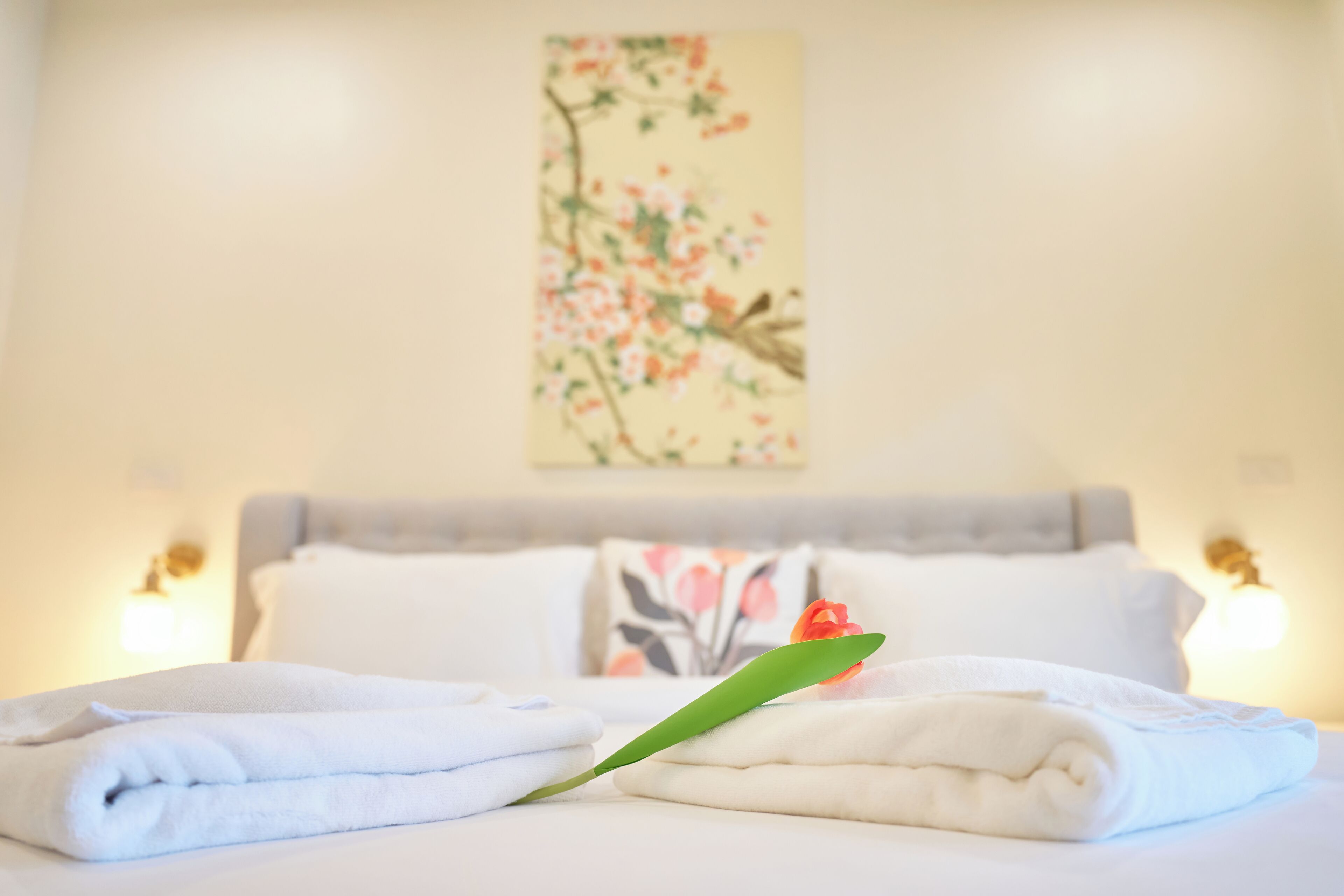 A neatly made bed with folded towels and a green leaf decoration, a floral painting on the wall, and two wall-mounted lamps.