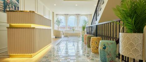 A hotel lobby with a reception desk, patterned floor tiles, a staircase, and potted plants.