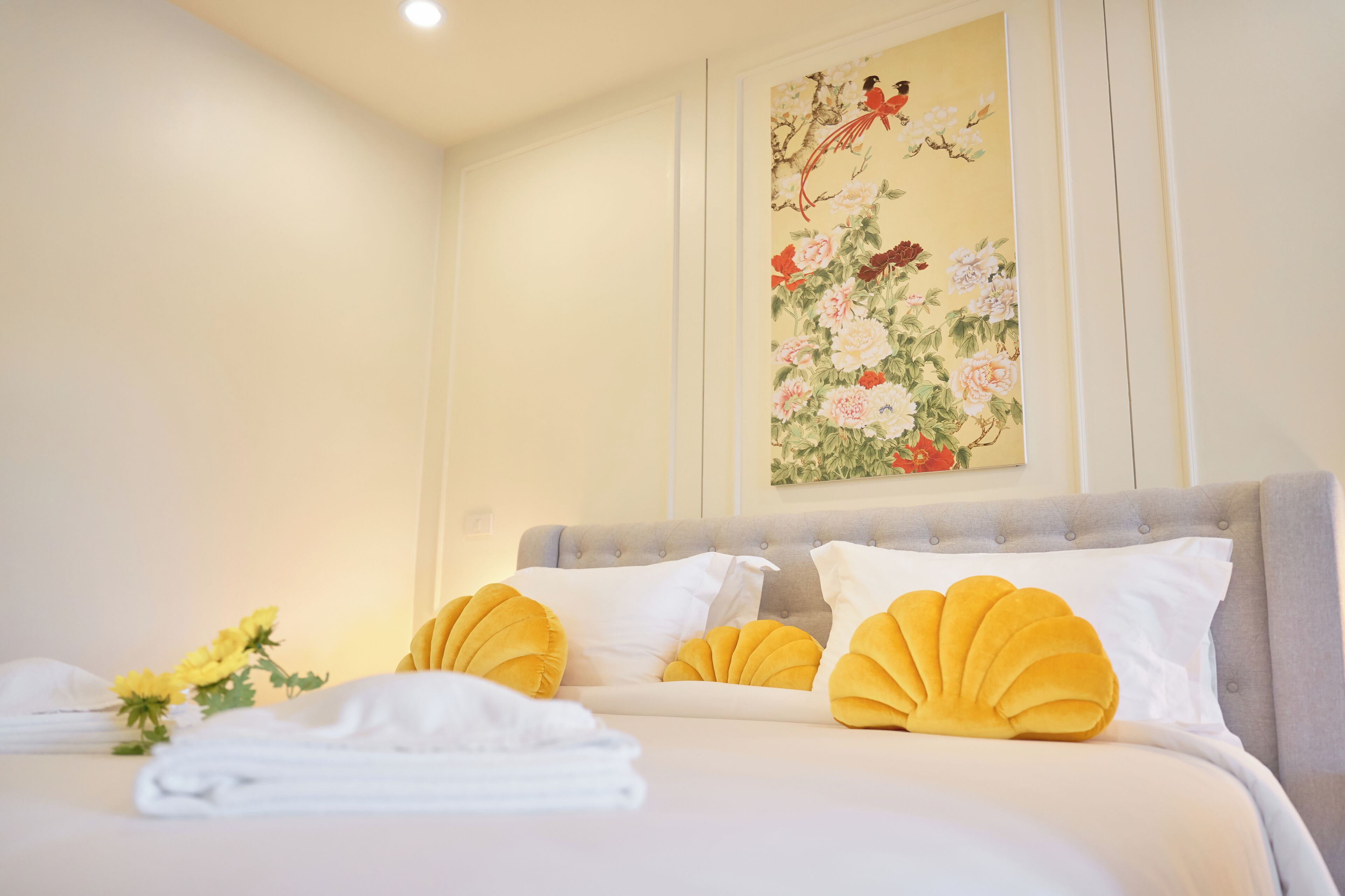 Una cama bien hecha con almohadas blancas y amarillas, una obra de arte floral en la pared y un ramo de flores amarillas.