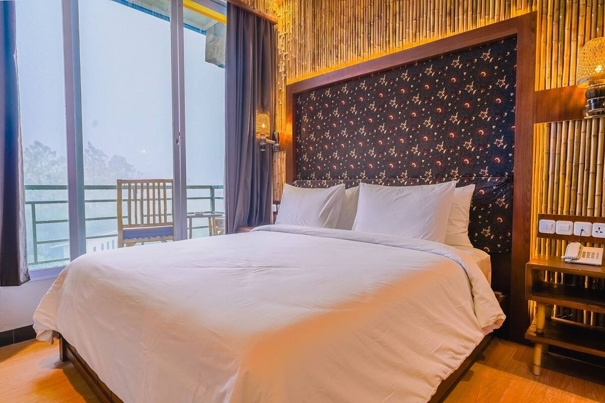A hotel room with a large bed, a wooden headboard with a star pattern, a bedside table with a lamp, and a view of the outdoors.