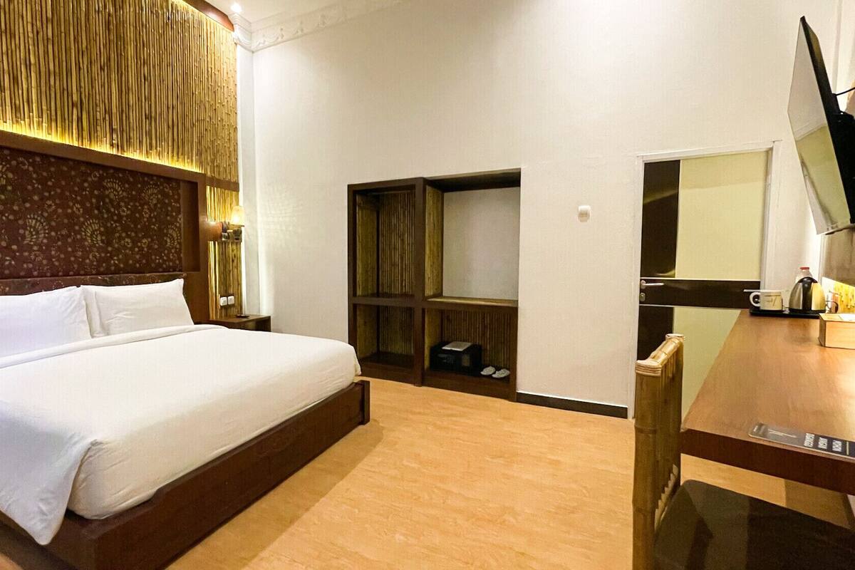 A hotel room with a large bed, a wooden headboard, a built-in wardrobe, a desk with a mirror, and a television mounted on the wall.