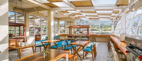 Un comedor amplio con mesas de madera y sillas azules, ventanas grandes que ofrecen vistas al exterior y un techo alto con luz natural.