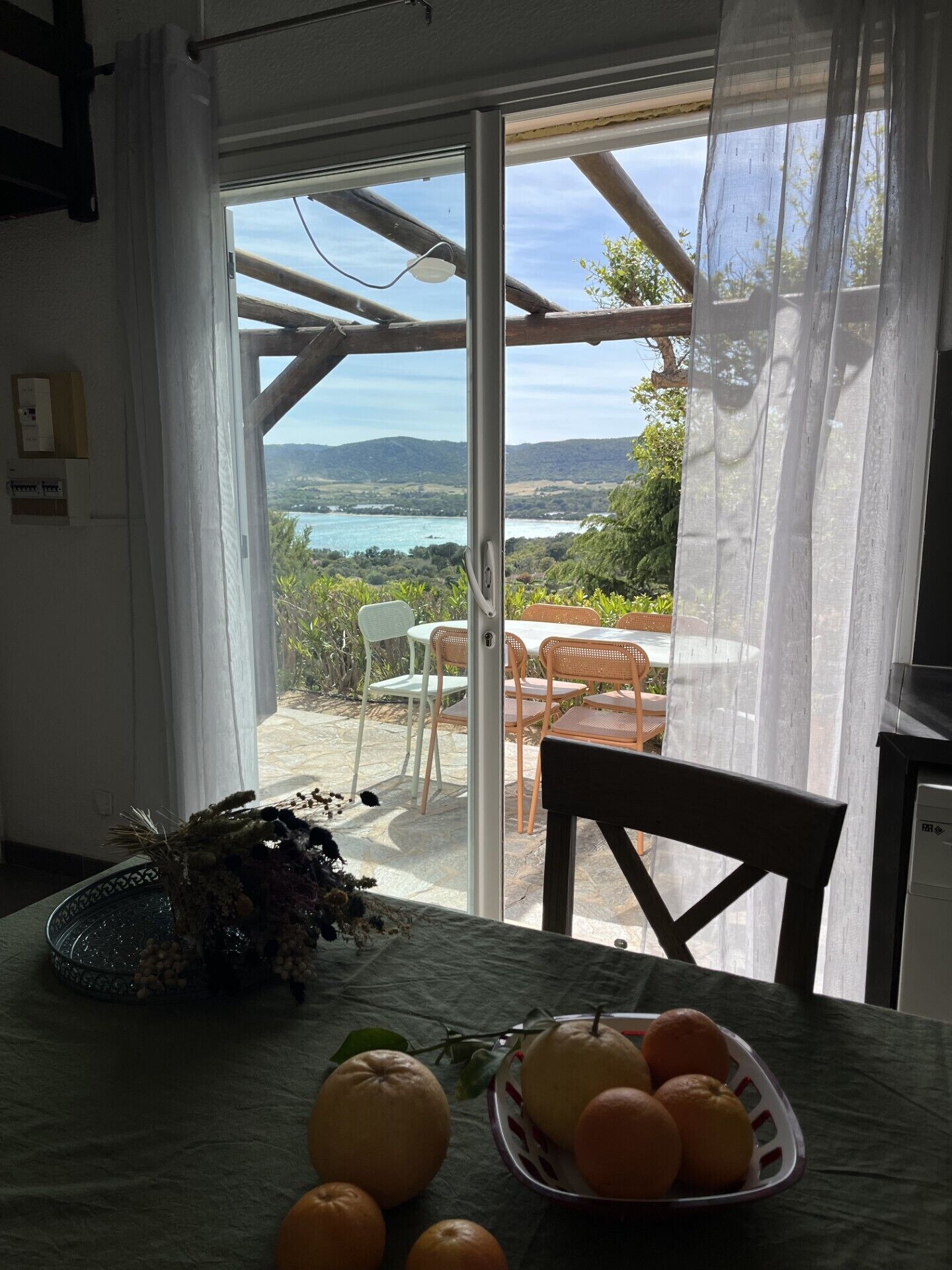 Un comedor con una mesa dispuesta para cuatro personas, una cesta de naranjas y vistas a un paisaje costero a través de un ventanal.