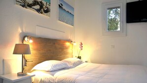 Un lit bien fait, avec du linge de lit blanc, une tête de lit en bois, une lampe de chevet et une petite table de chevet avec une lampe. Il y a une fenêtre donnant sur des arbres et un téléviseur fixé au mur.