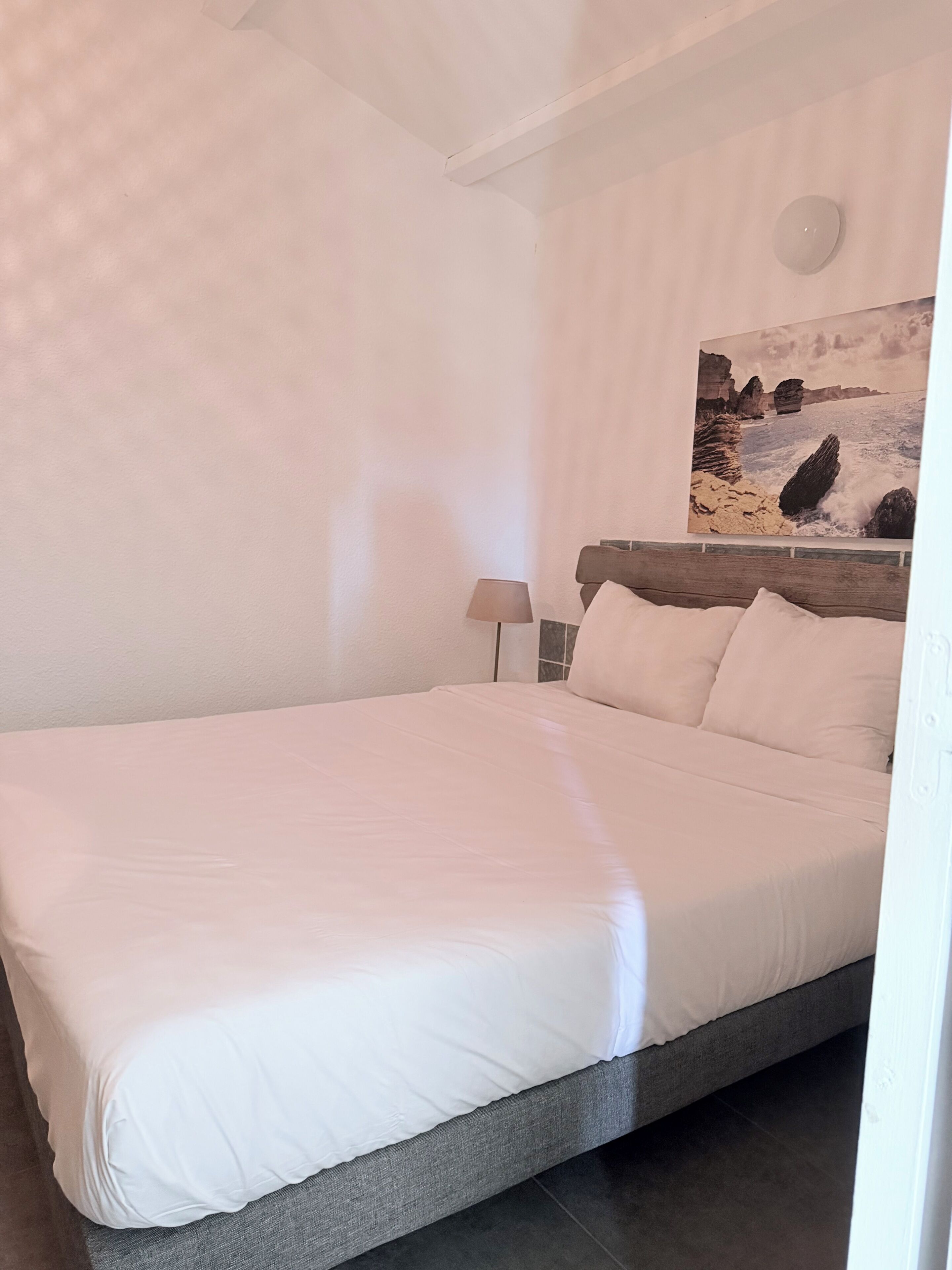A bedroom with a bed, a nightstand, a lamp, and a painting of a coastal scene.