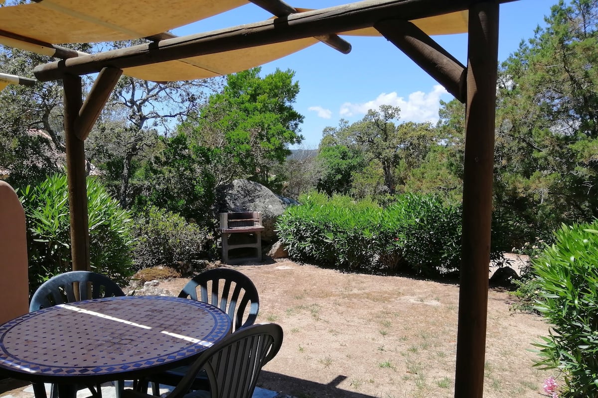 Een overdekt terras met een tafel en stoelen, omringd door groen en een rotsformatie.