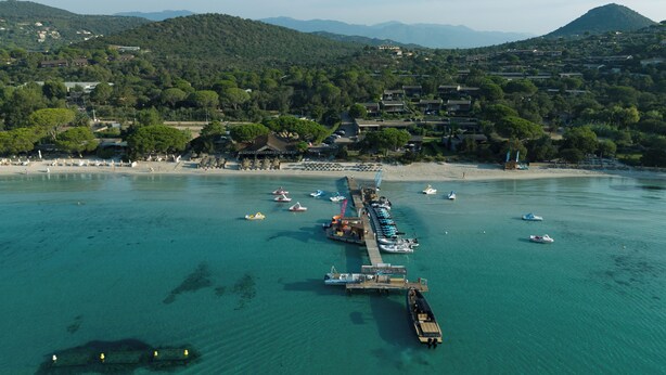 Vue aérienne d’une zone côtière avec une jetée s’avançant dans des eaux turquoise, entourée d’une végétation luxuriante et de bâtiments résidentiels.