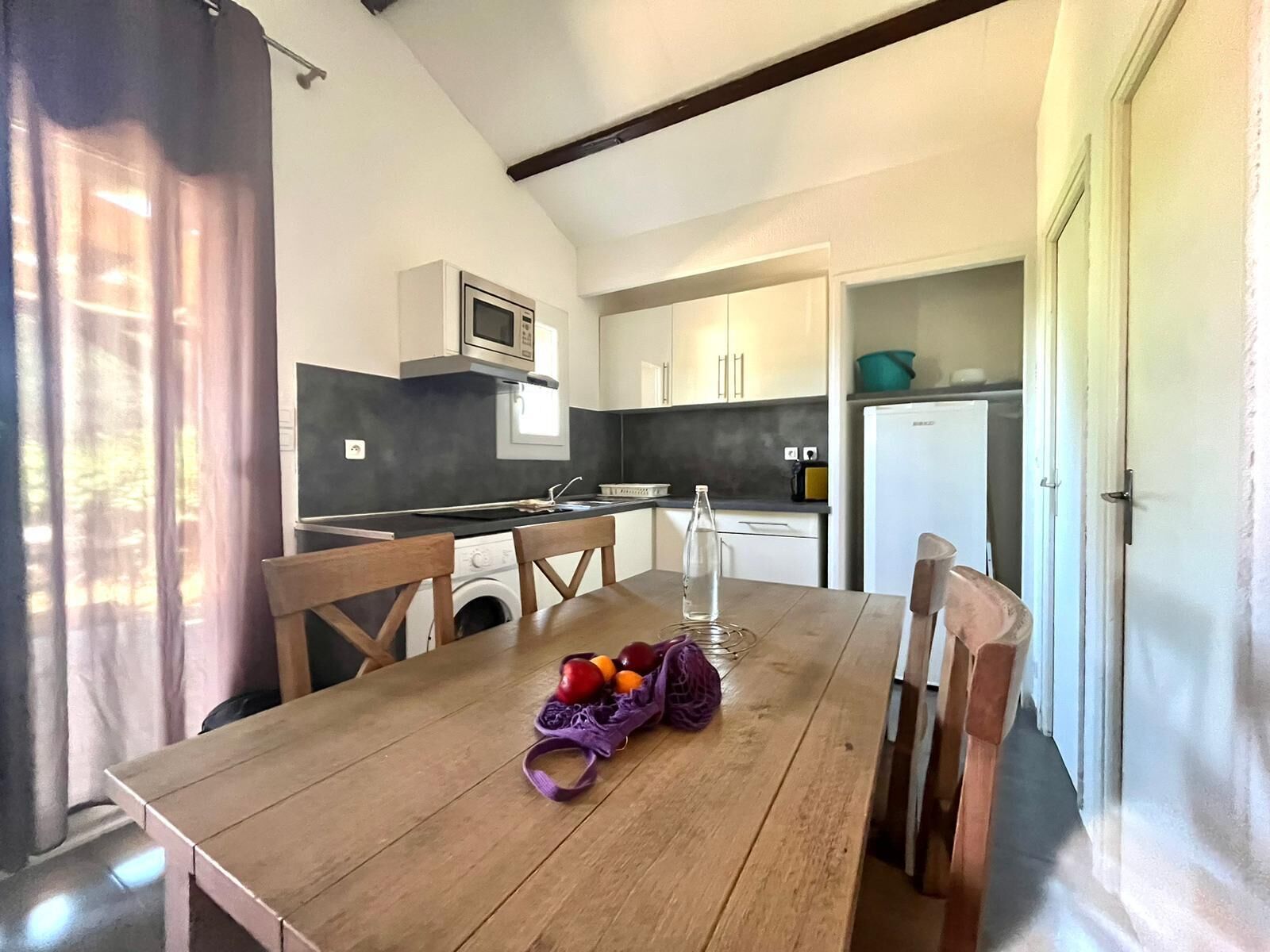 A compact kitchen with a microwave, sink, and a small refrigerator. There is a wooden table with chairs and a basket of fruit.