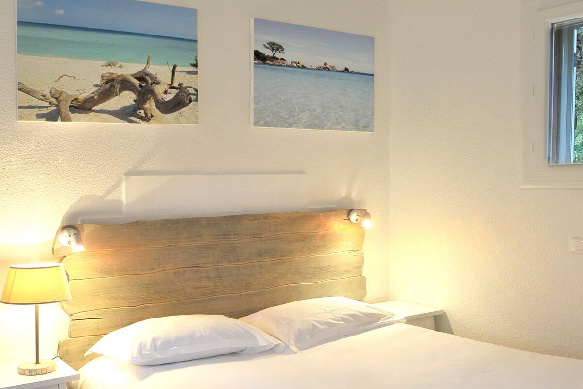 Een net opgemaakt bed met witte lakens, een houten hoofdbord en een nachtkastje met een lamp. Boven het bed hangen twee ingelijste foto's aan de muur.