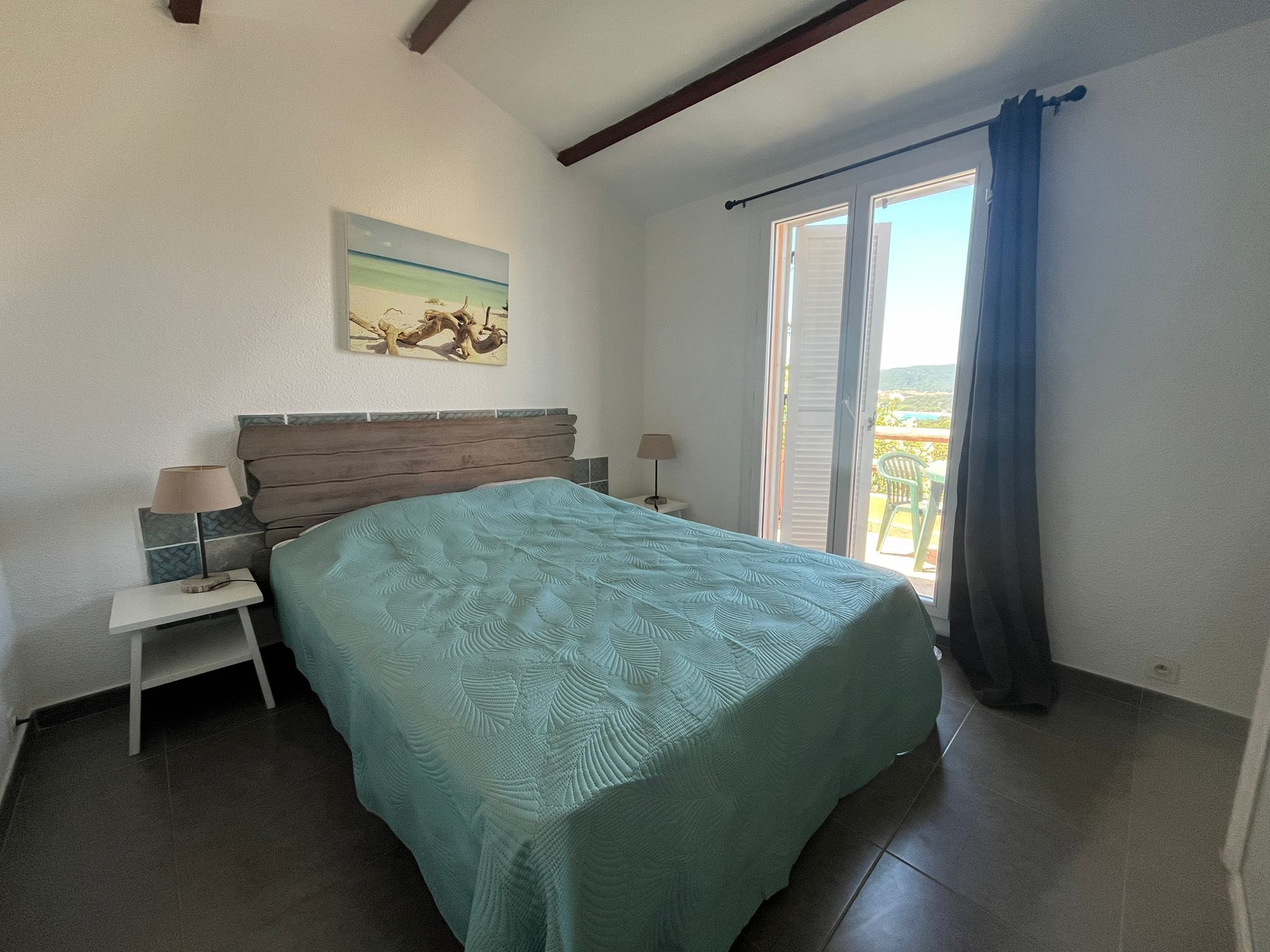Un dormitori amb un llit, una tauleta de nit, una làmpada, un quadre a la paret i una vista per la finestra.