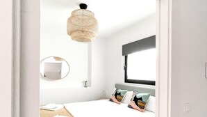 Ein modernes Schlafzimmer mit Bett, rundem Spiegel, Pendelleuchte und Fenster mit schwarzem Rollo.
