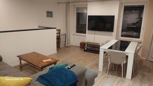 Un salon avec une table basse en bois, un canapé, une petite table blanche avec une chaise, et un téléviseur fixé au mur.