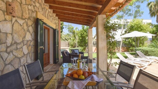 A covered outdoor dining area with stone walls, wooden beams, and a glass table with a fruit basket.