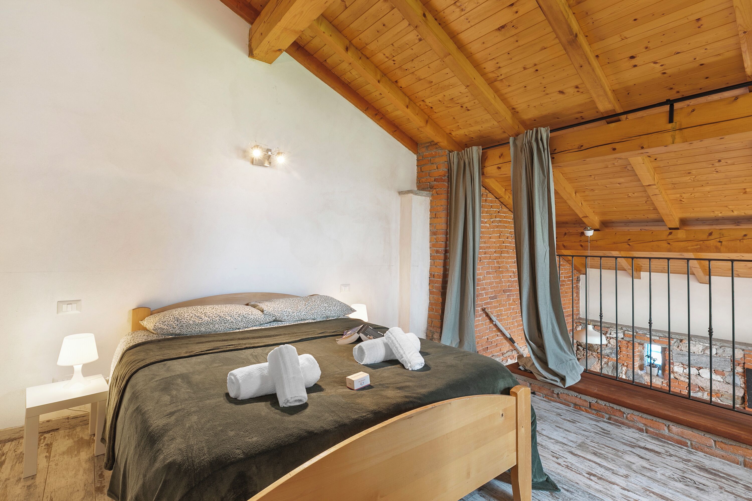 Un dormitorio con cama de madera, una mesita de noche, pared de ladrillo y vistas desde un balcón.