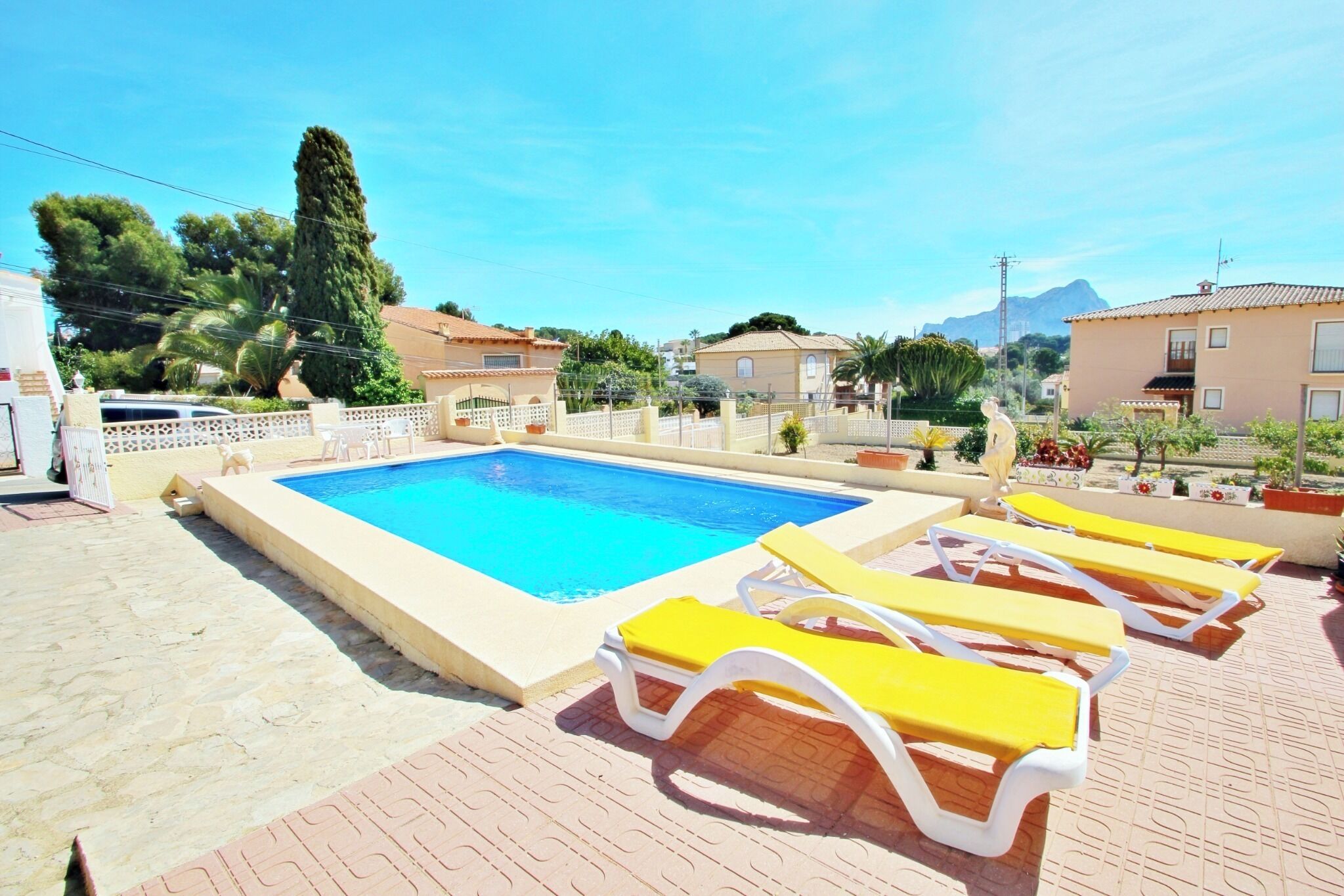 A swimming pool with sun loungers, surrounded by a fence and palm trees.