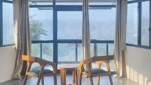 Una habitación con cama, dos sillas de madera, una mesita y ventanas grandes con cortinas.