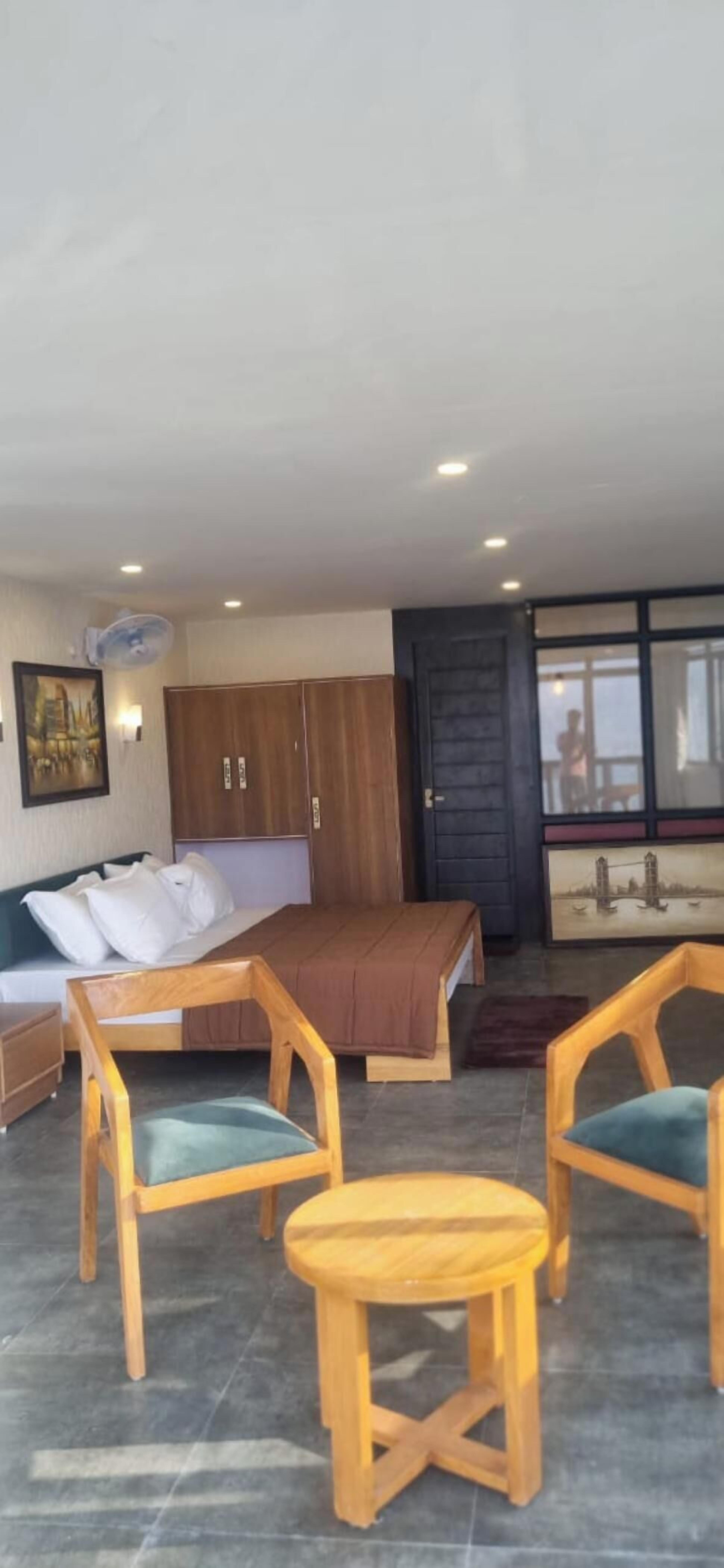 Quarto de hotel com uma cama, cadeiras de madeira, uma pequena mesa, um ventilador e um quadro na parede.