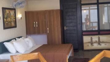 Quarto de hotel com uma cama, cadeiras de madeira, uma pequena mesa, um ventilador e um quadro na parede.