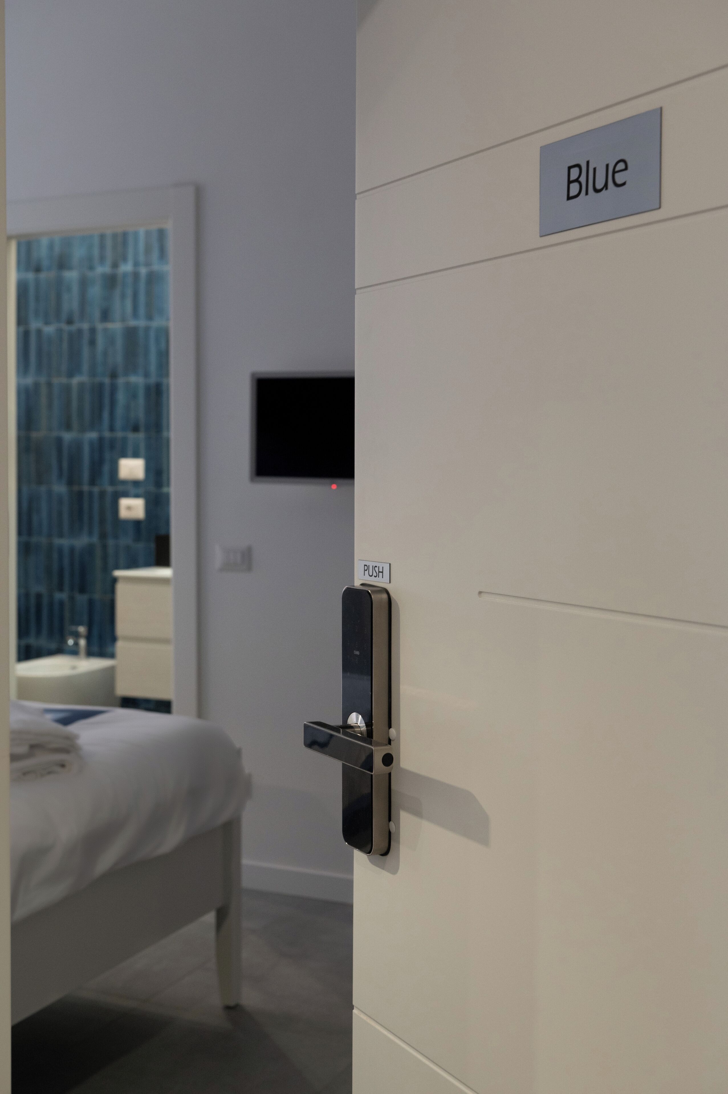 A hotel room with a bed, a bathroom with a blue tiled wall, a television mounted on the wall, and a door labeled "Blue".