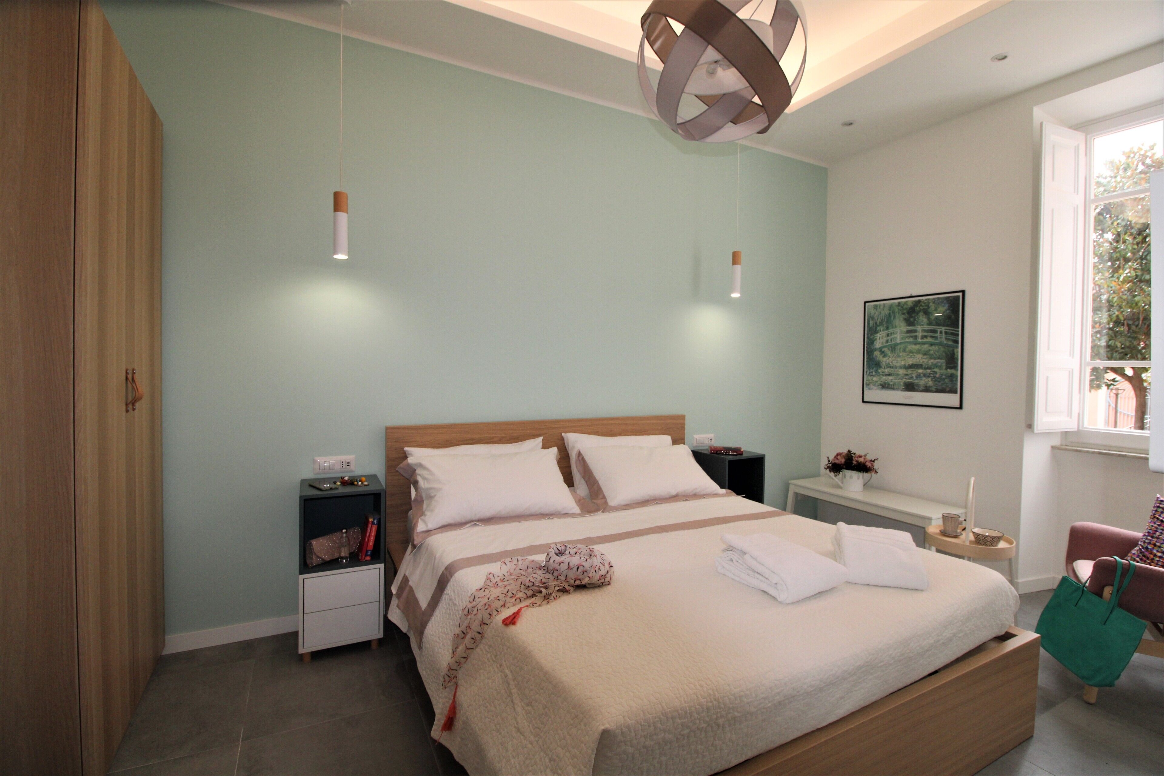 Een net opgemaakt bed met witte lakens, een nachtkastje met een lamp en een raam met uitzicht op bomen.