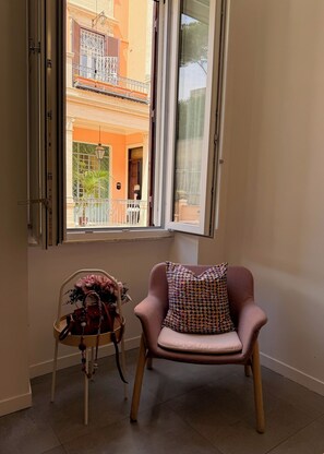 A room with a chair and a small table, a window with an open shutter, and a view of a building with balconies.