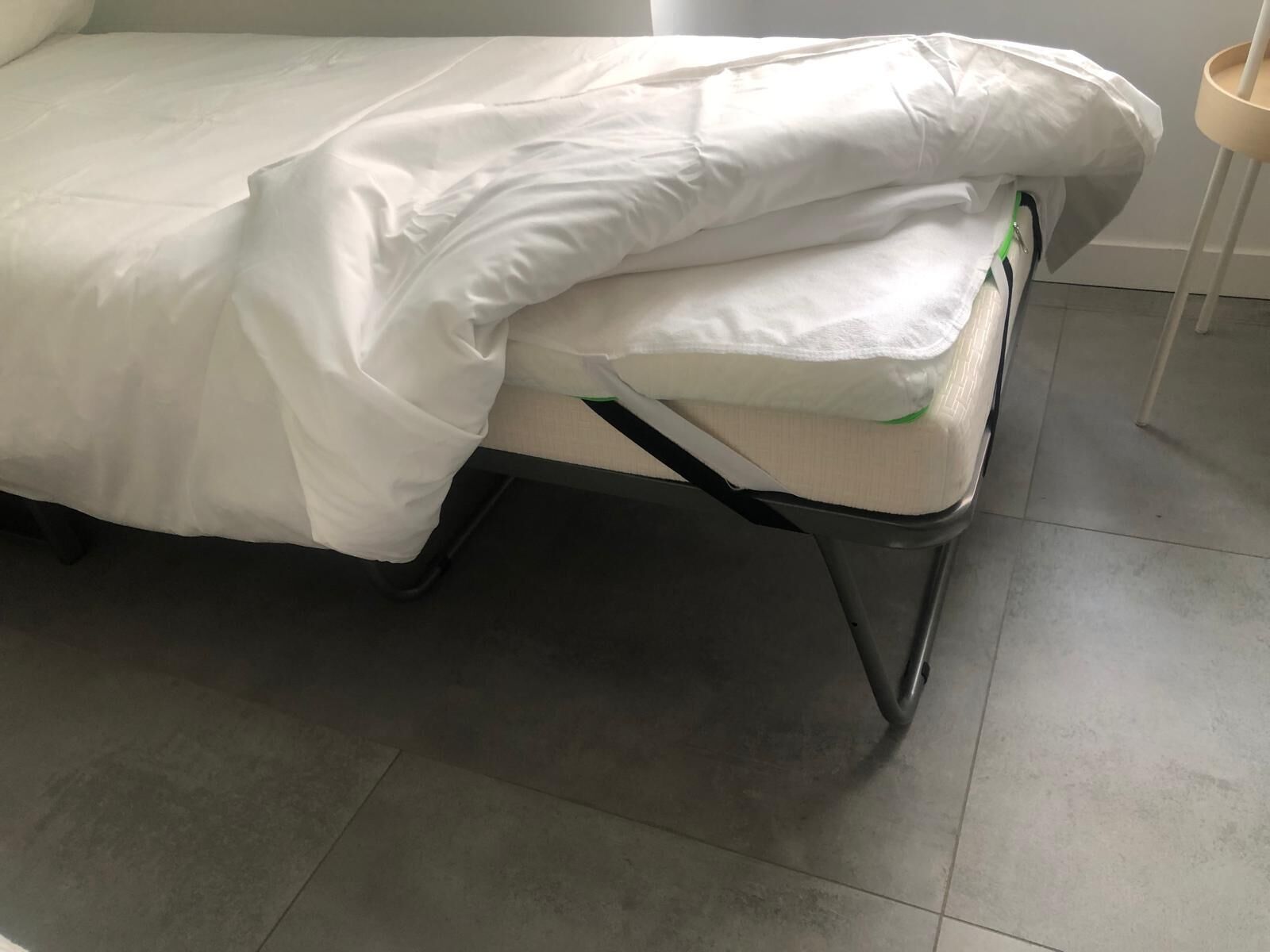 A bed with a white mattress and a white cover, a metal frame, and a grey tiled floor.