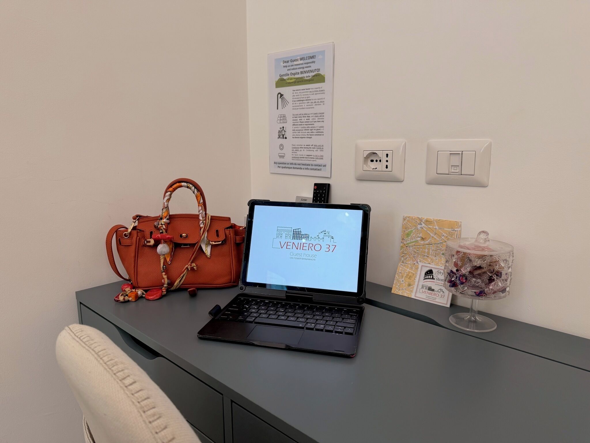 A laptop on a desk with a sign above it, a remote control, a glass jar with flowers, and a handbag.