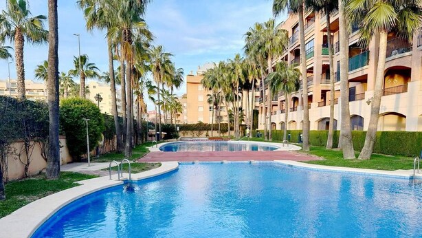 A swimming pool with a curved edge, surrounded by palm trees and a multi-story building with balconies. - Beautiful and comfortable apartment in Denia, Costa Blanca, Spain  with communal pool for 4 persons (Denia)