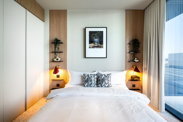 A modern hotel room with a large bed, two bedside lamps, a framed picture on the wall, and a window with curtains.