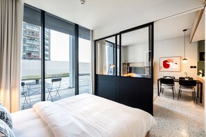 A modern hotel room with a large bed, a dining area, and a view of a building through large windows.