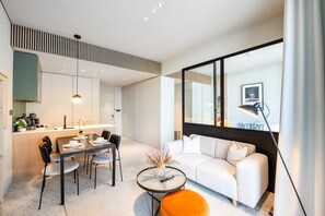 A modern living room with a white sofa, a dining table with chairs, a coffee machine, and a large window with a view.