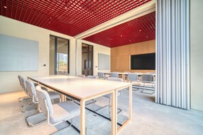 A conference room with a long table, grey chairs, a red ceiling, and a large screen.