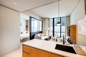 A modern hotel room with a large bed, a sitting area with a sofa and chairs, a dining table with a chandelier, and a view of the city through large windows.