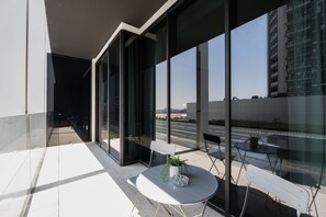 An outdoor balcony with a table and chairs, large windows, and a view of a building in the background. - Modern 1BR w/ Balcony in Business Bay (Dubai)