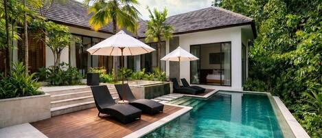 A modern pool area with lounge chairs and umbrellas, surrounded by lush greenery and a building with a tiled roof.