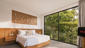 A modern bedroom with a large bed, a decorative wall panel, a bedside table with a lamp, and a view of greenery through a sliding glass door. - Villa Talun by GORO (Ubud)