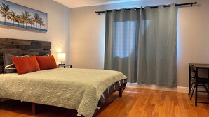 Un dormitorio con cama, cabecera de madera, mesita de noche con lámpara, un cuadro de palmeras y una ventana con cortinas.