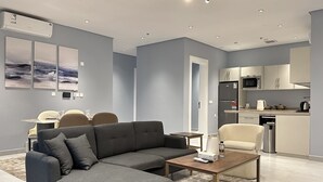 Ein modernes Wohnzimmer mit grauem Sofa, einem Couchtisch und einem Essbereich mit Tisch und Stühlen. Im Hintergrund ist eine Küche mit Mikrowelle und Kühlschrank zu sehen.
