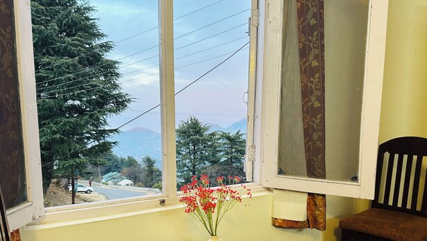 A dining area with a table set for two, a vase with red flowers, and a view of a road and trees outside. - Hotel Eagle Nest Central Heated  (Dalhousie)
