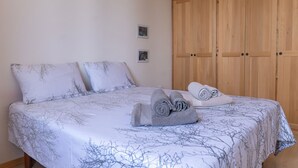 Una cama bien hecha con una colcha estampada con motivos de ramas de árbol, dos almohadas y toallas enrolladas sobre un suelo de madera.